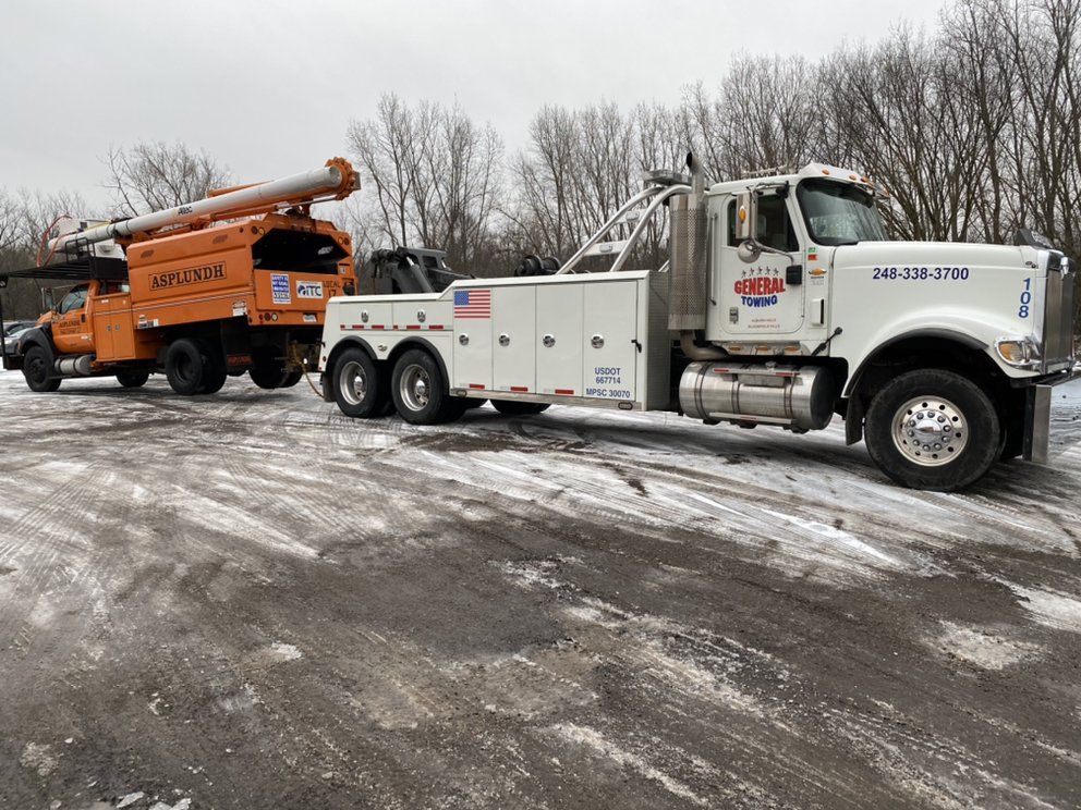 White tow truck towing an orange tree trimmer on a snowy lot.