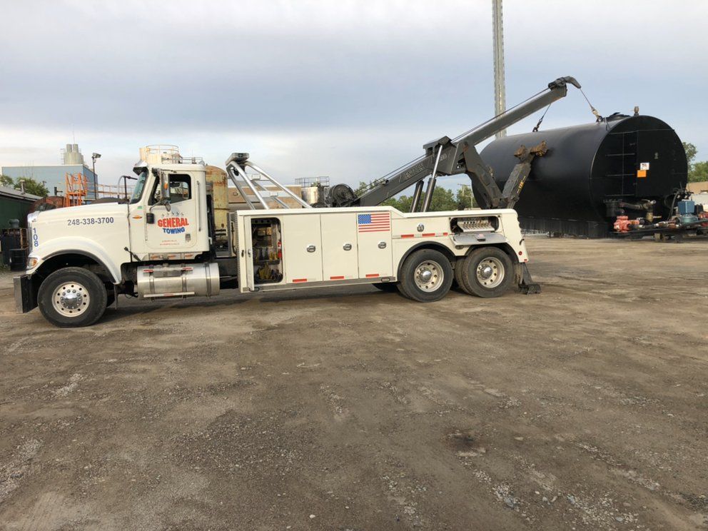 White tow truck lifting a large black tank in a gravel lot.