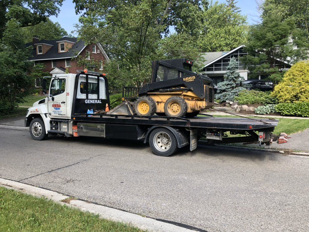 Tow truck with a yellow skid steer loader parked on a residential street.