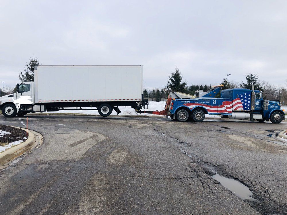 Tow truck towing a white box truck on a cracked asphalt road in a snowy setting.