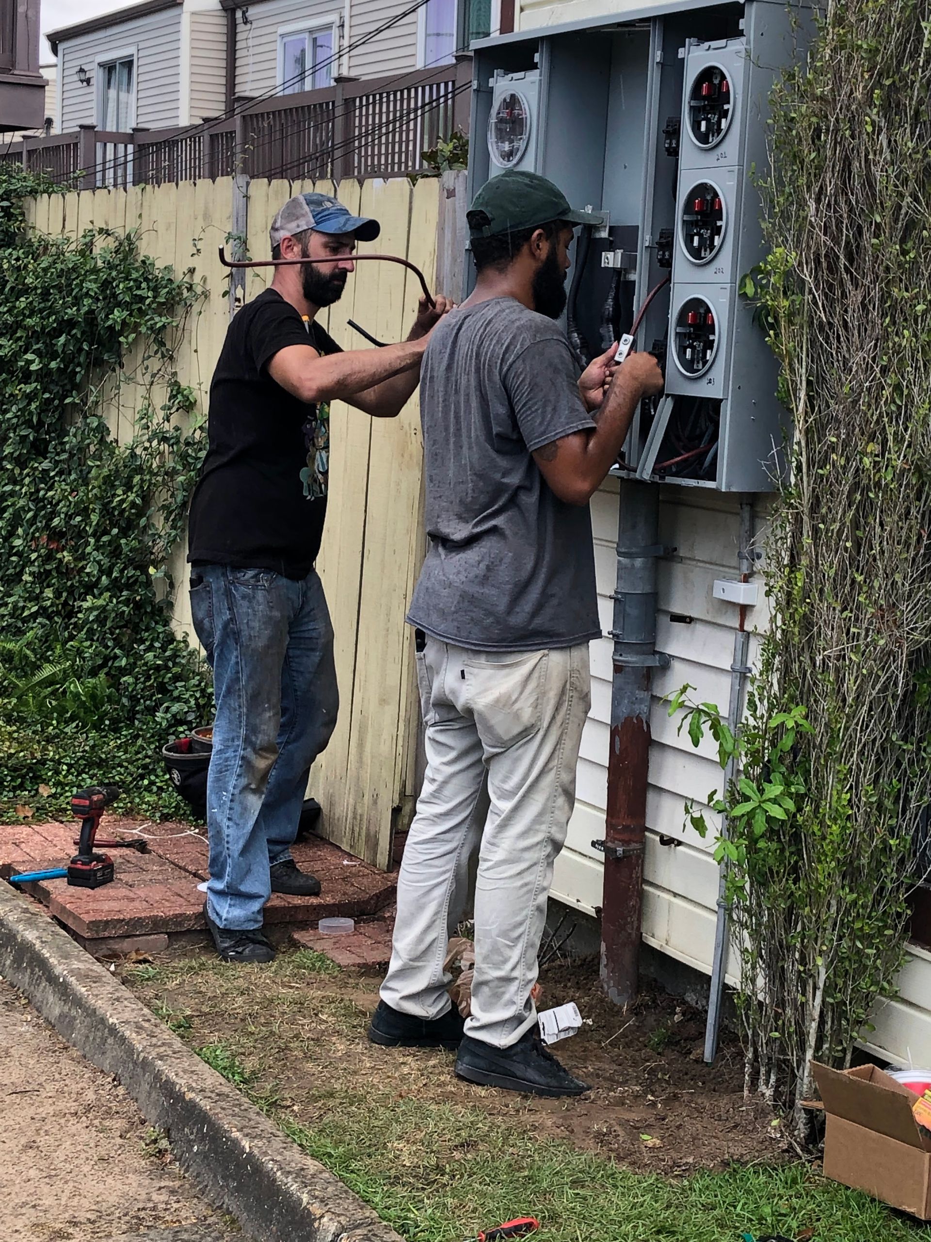 Two men are working on an electrical box on the side of a house.