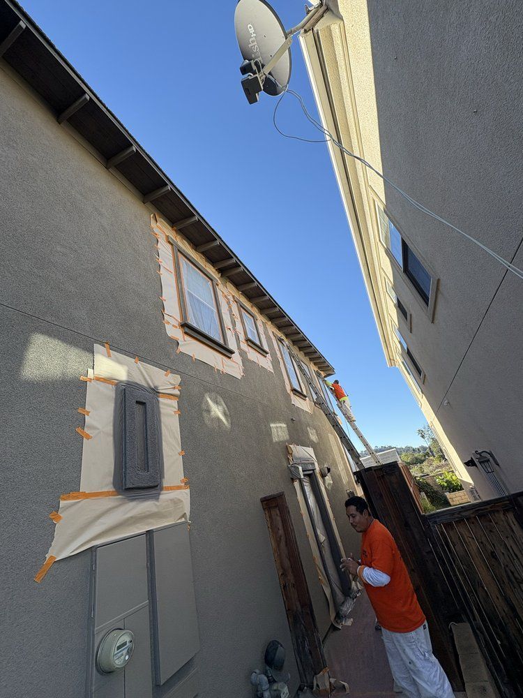A person in orange painting the exterior wall of a house with a ladder. Blue sky in the background.