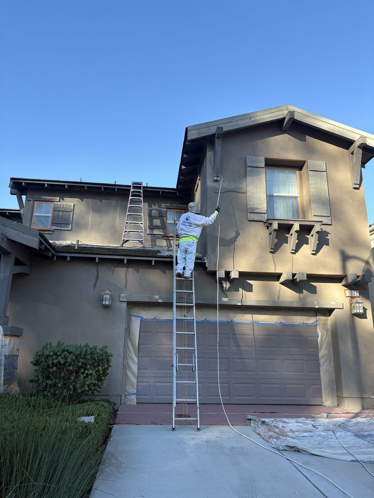 Person in protective suit painting a two-story house, standing on a ladder. House has a garage door and brown shutters.