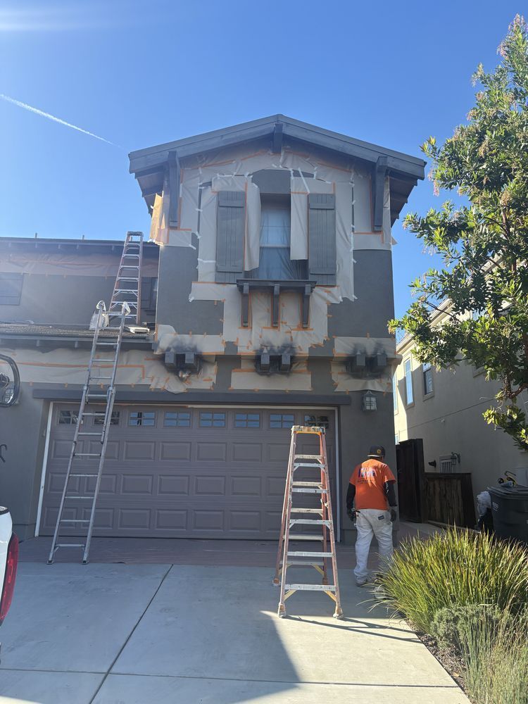 House exterior being painted; workers on ladders. Garage, blue sky, and a tree are also visible.