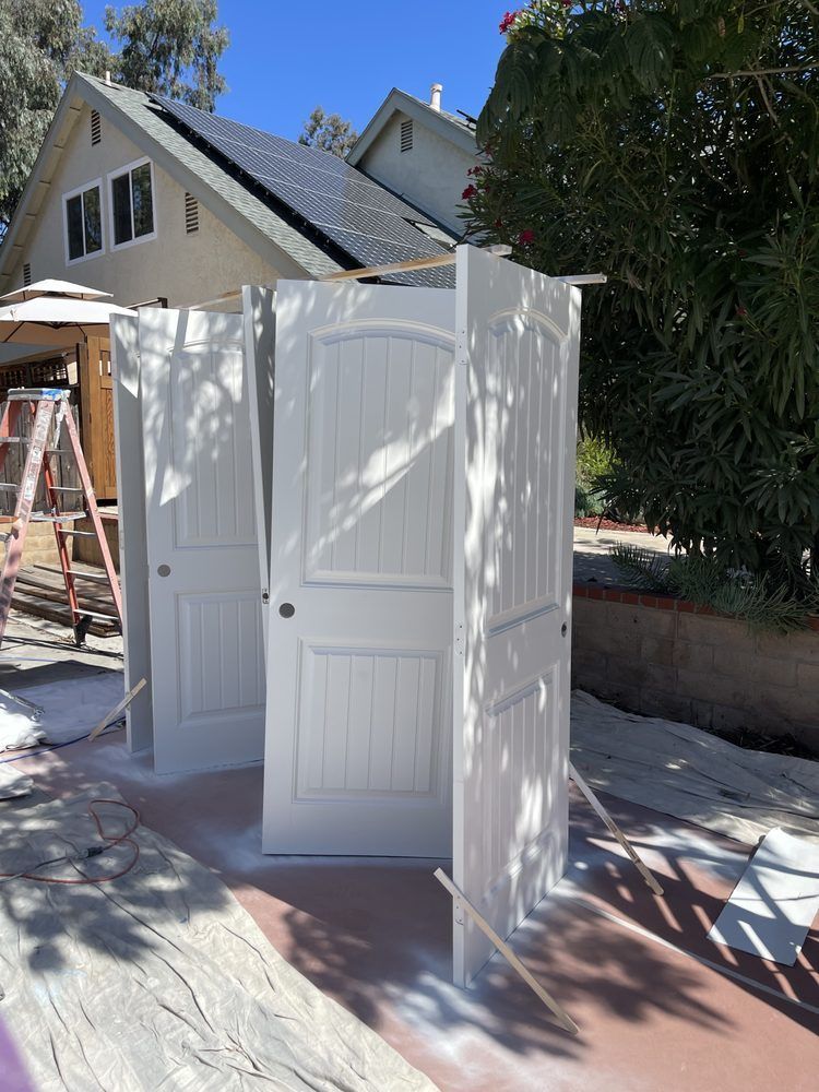 Three white doors propped up outside, being spray painted in front of a house on a sunny day.