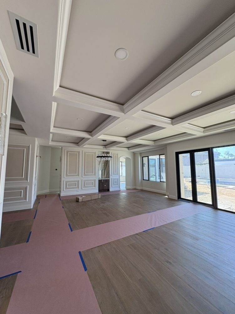 An empty room with light wood floors, coffered ceiling, large windows, and decorative wall panels.