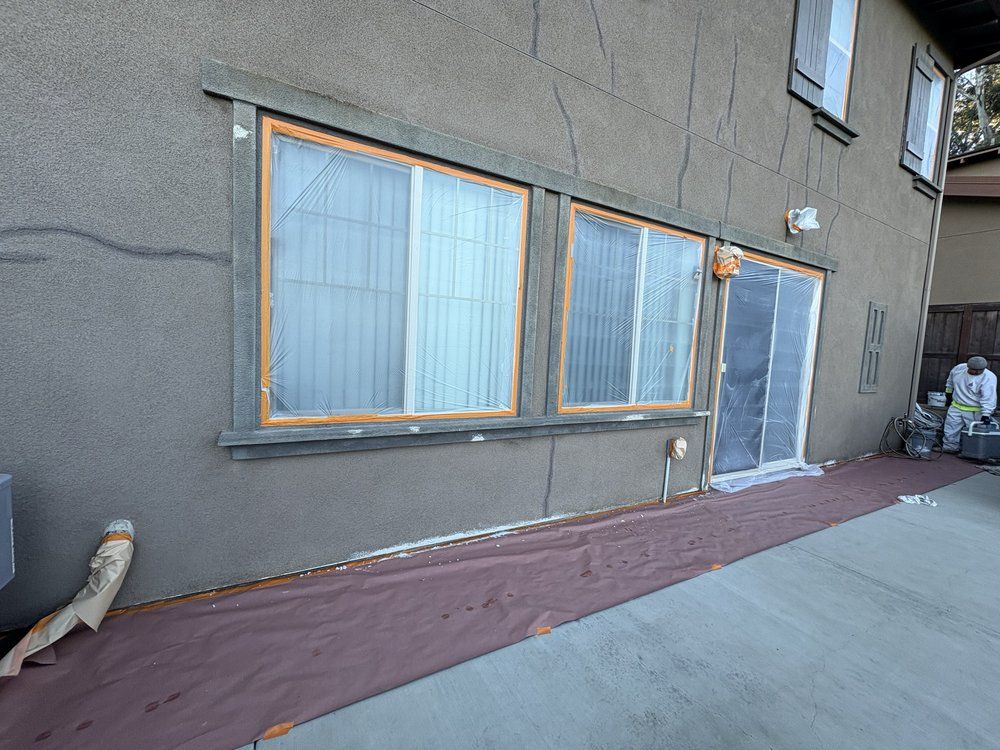 Exterior view of a building with windows and a sliding door. The exterior is gray, with a protective tarp laid out.