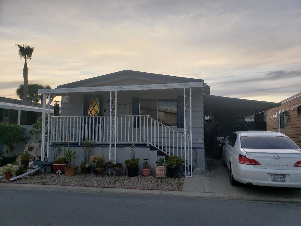 Mobile home with carport and white car parked in front; porch with plants.