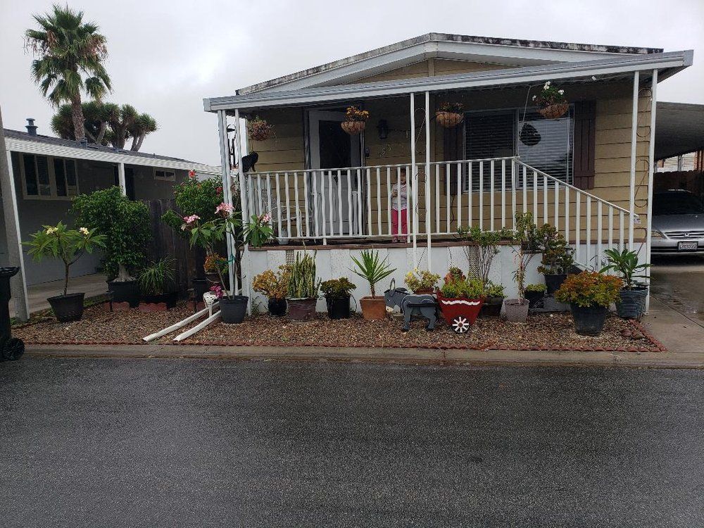 Mobile home with a porch, plants, and a cloudy sky.