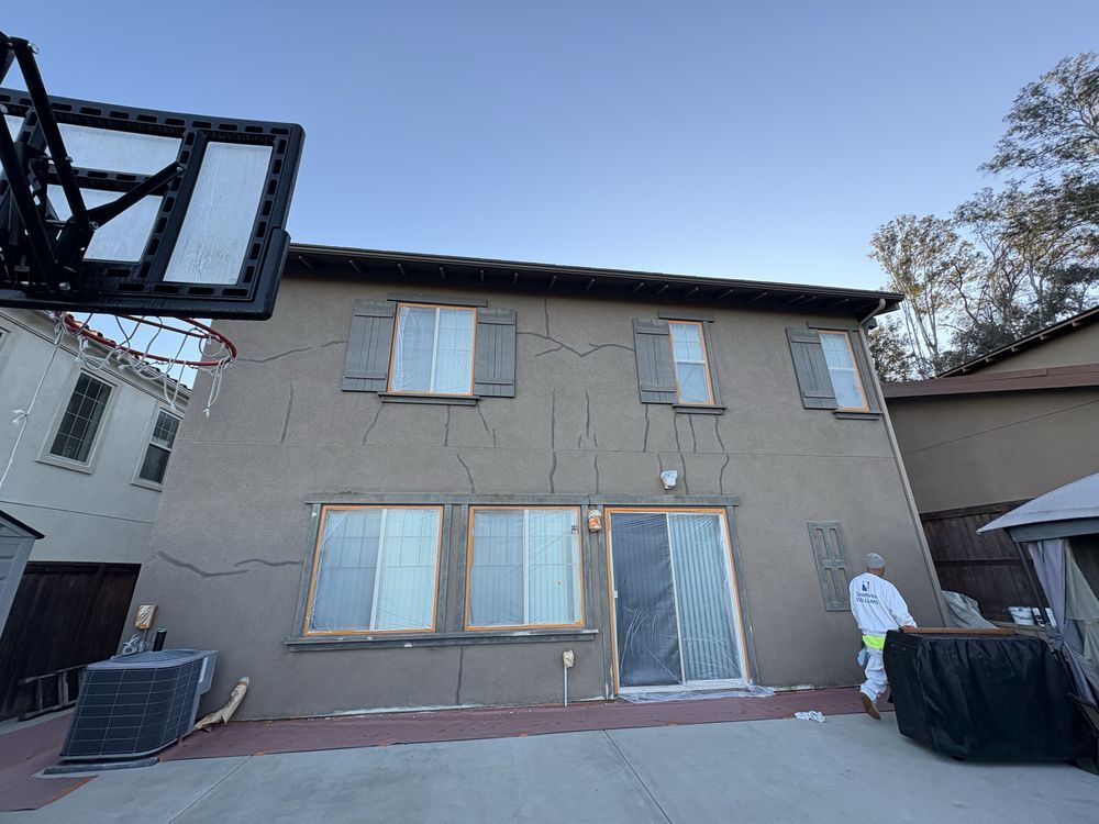 Back of a two-story stucco house with visible cracks. A person in a hazmat suit works on the side. Basketball hoop in view.