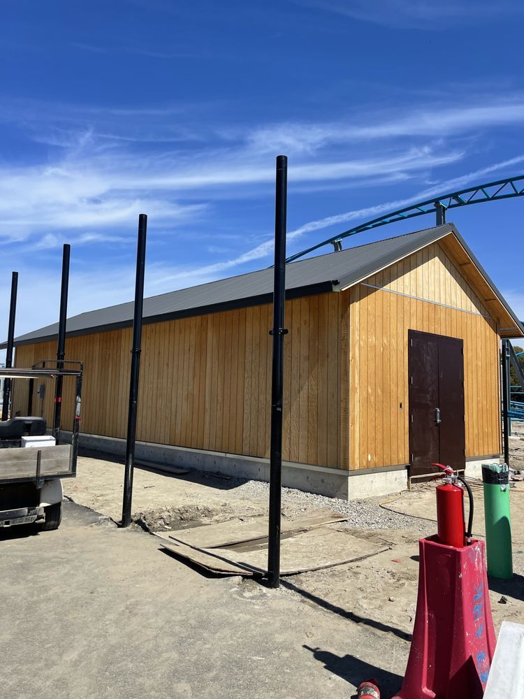 Wooden building under construction, with a gray roof and brown door. Black poles stand in front. Blue sky.