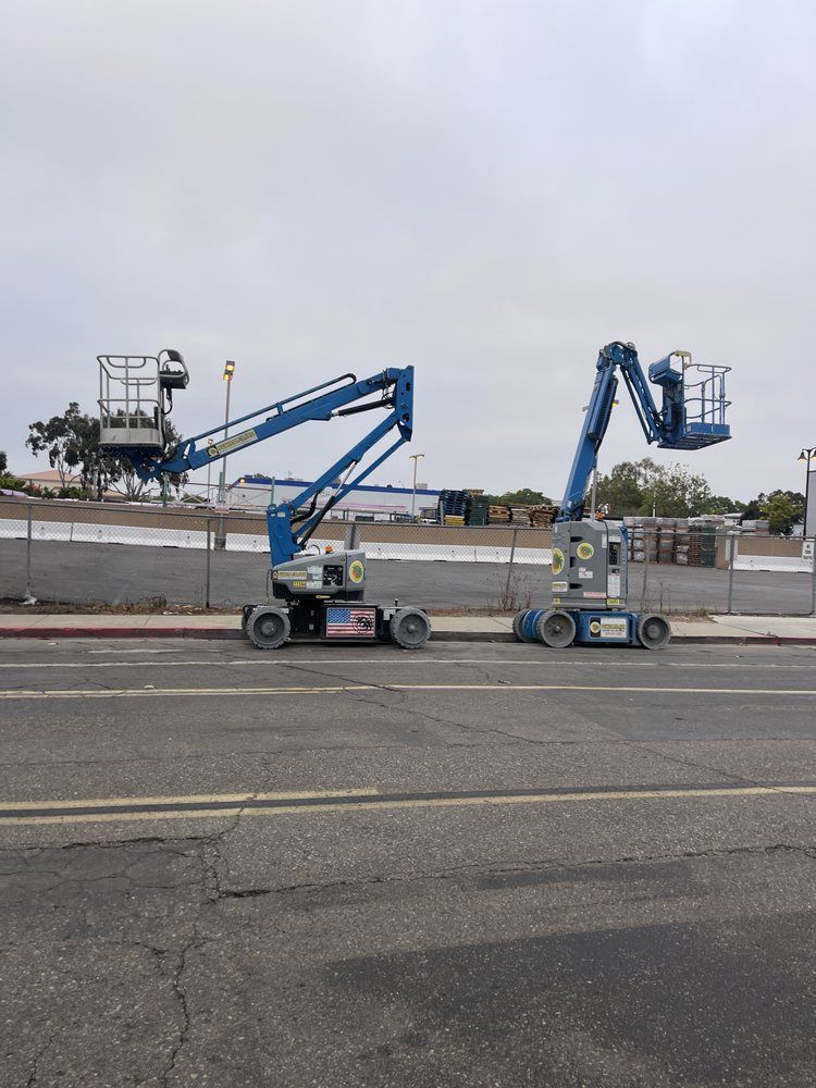 Two blue articulated boom lifts parked on a sidewalk next to a road, construction site in background.