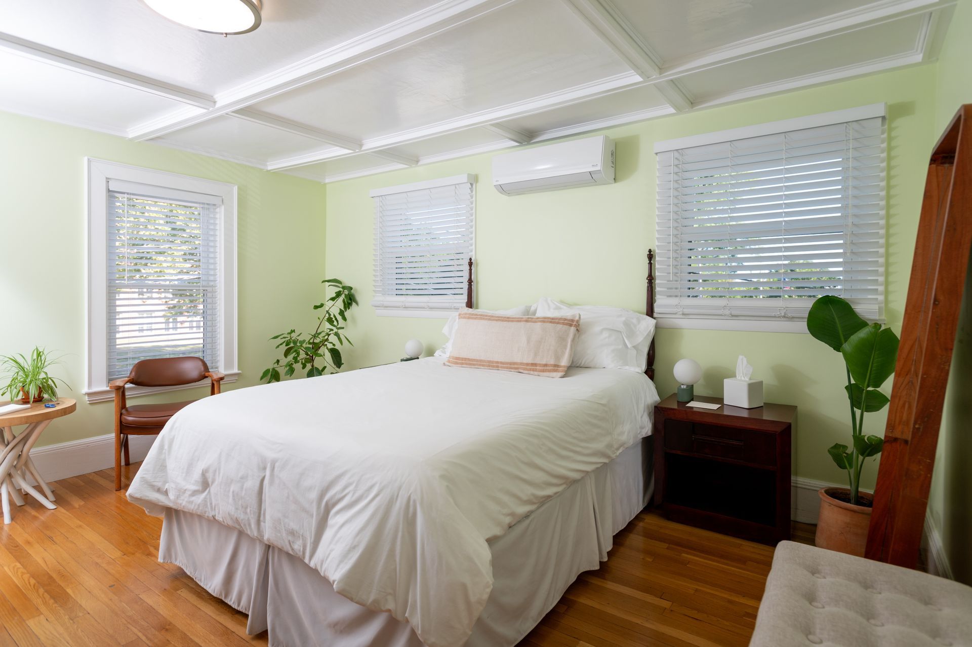 Bedroom with bed, windows, and plants; light green walls, wood floor, and decorative ceiling.