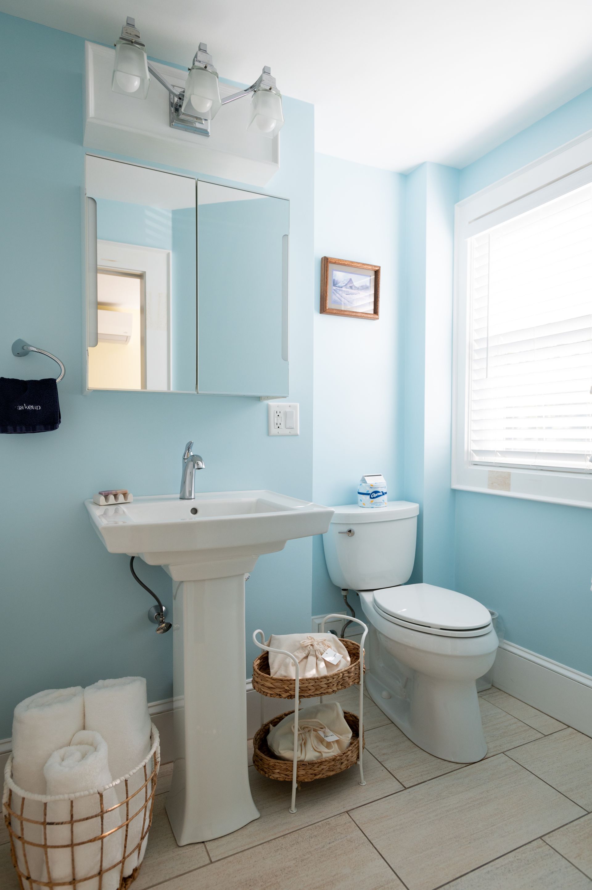 Blue bathroom with pedestal sink, toilet, and white framed window.