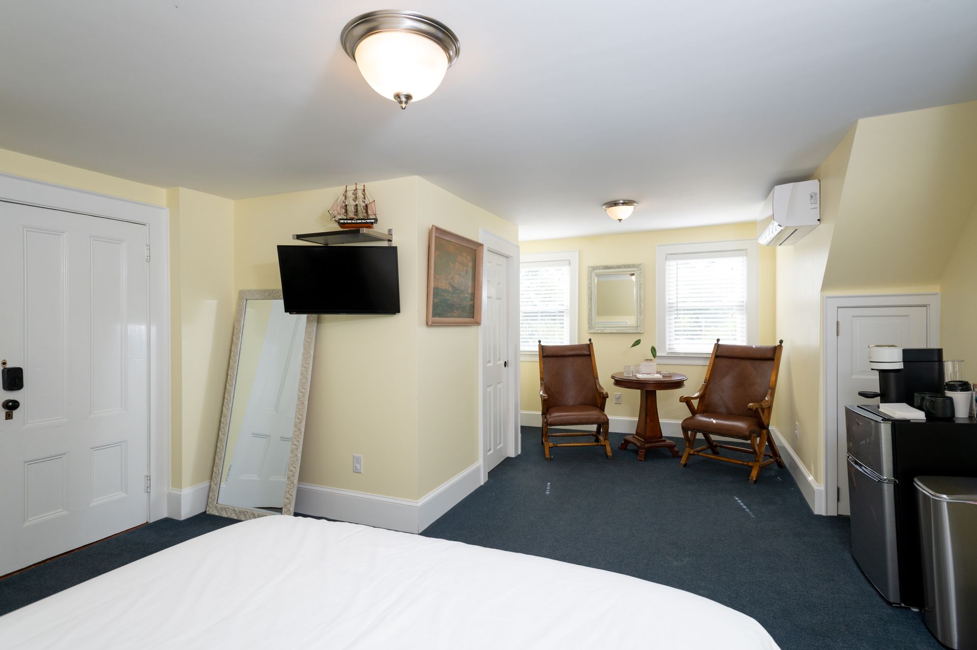 Bedroom with bed, chairs, TV, and mini-fridge in a room with yellow walls and a blue carpet.