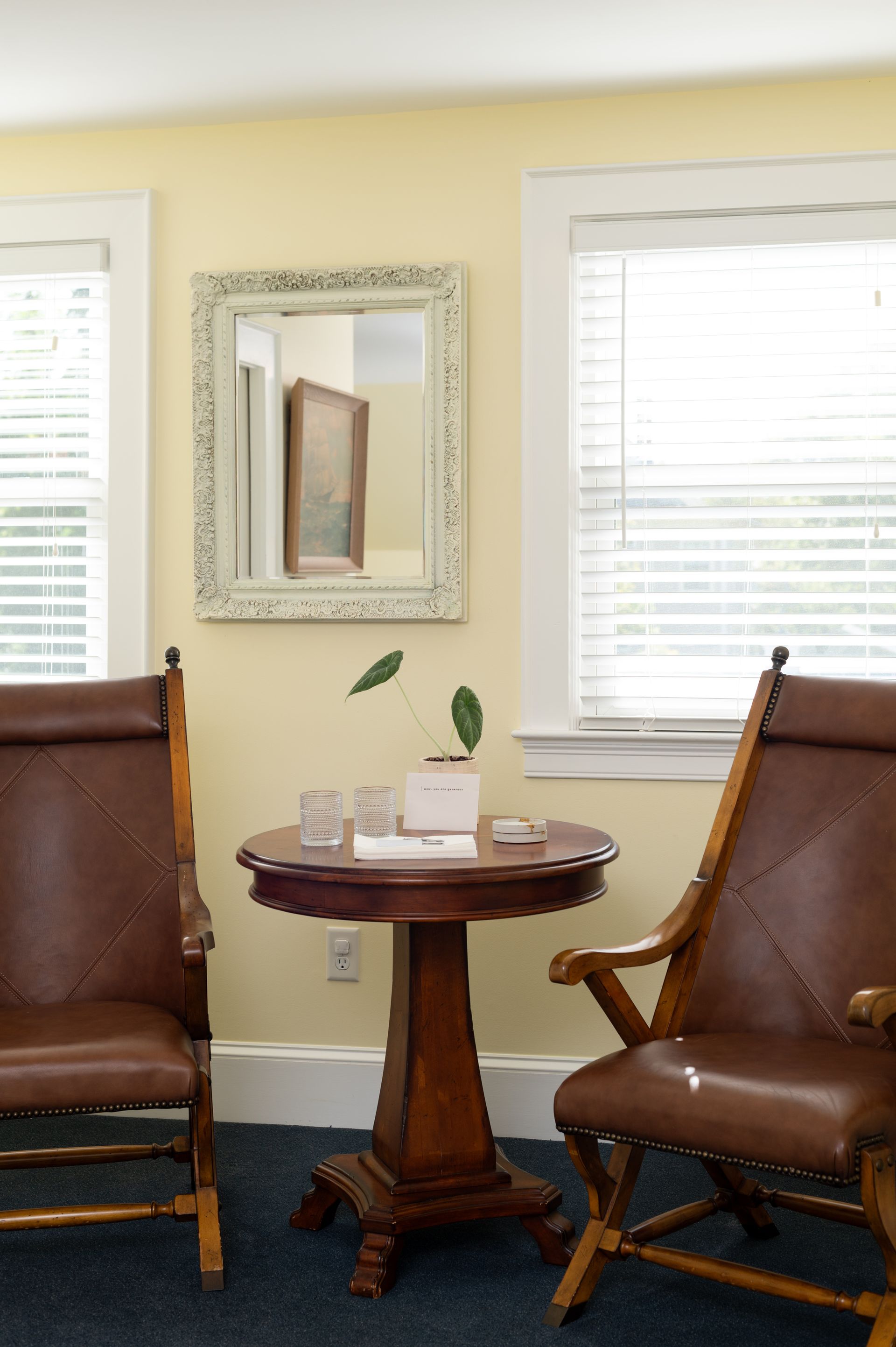 Two brown leather chairs flank a small table with a mirror above it, near windows.