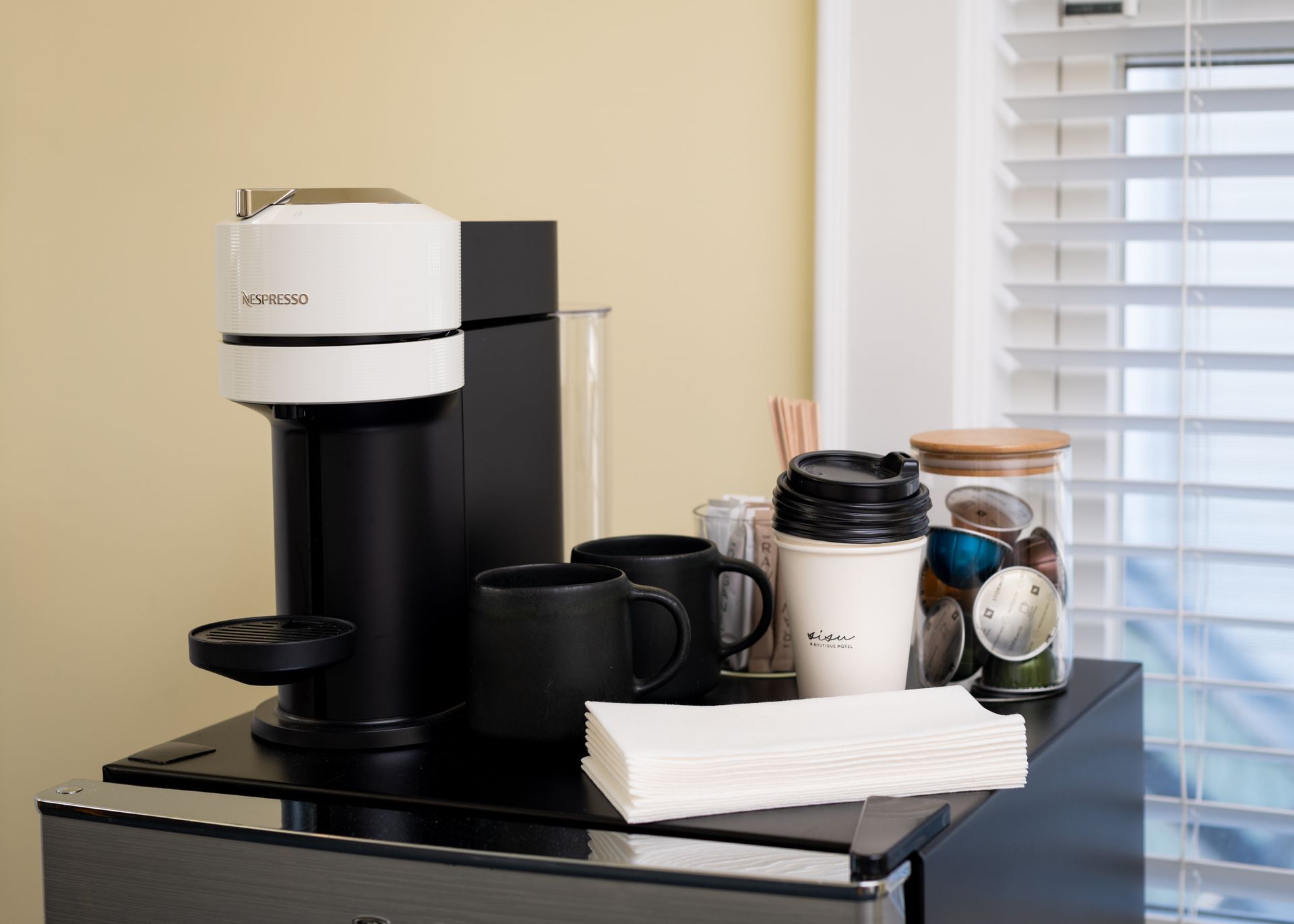 Coffee machine, mugs, and supplies on a counter near a window with blinds.