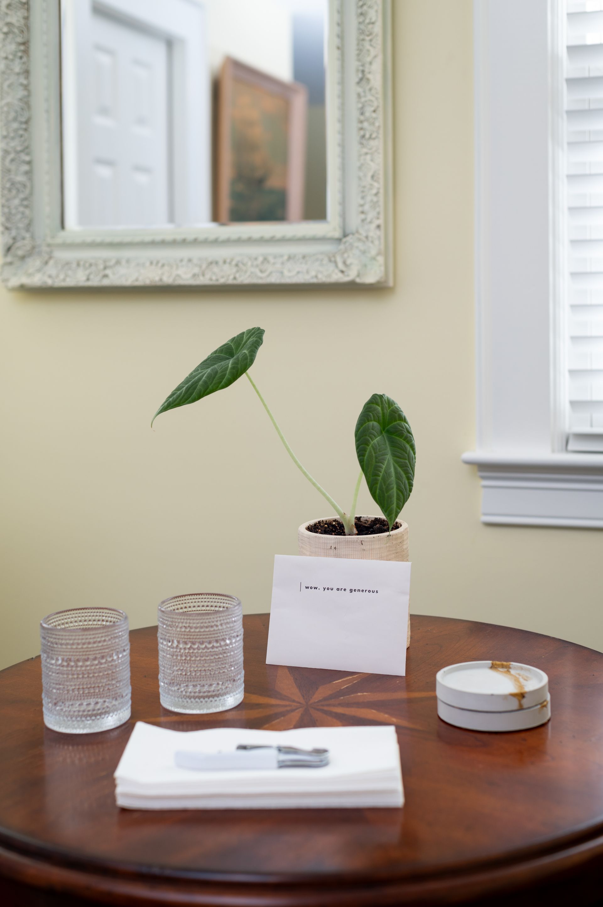 Round table with a plant, glasses, card, and box in front of a mirror and window.