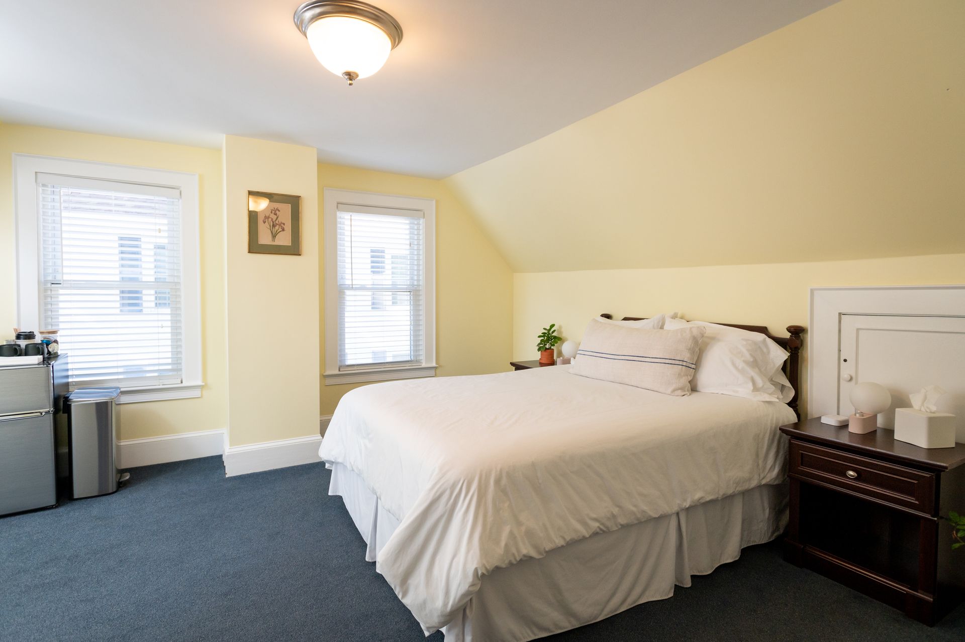 Bedroom with bed, windows, and bedside table; yellow walls, blue carpet.