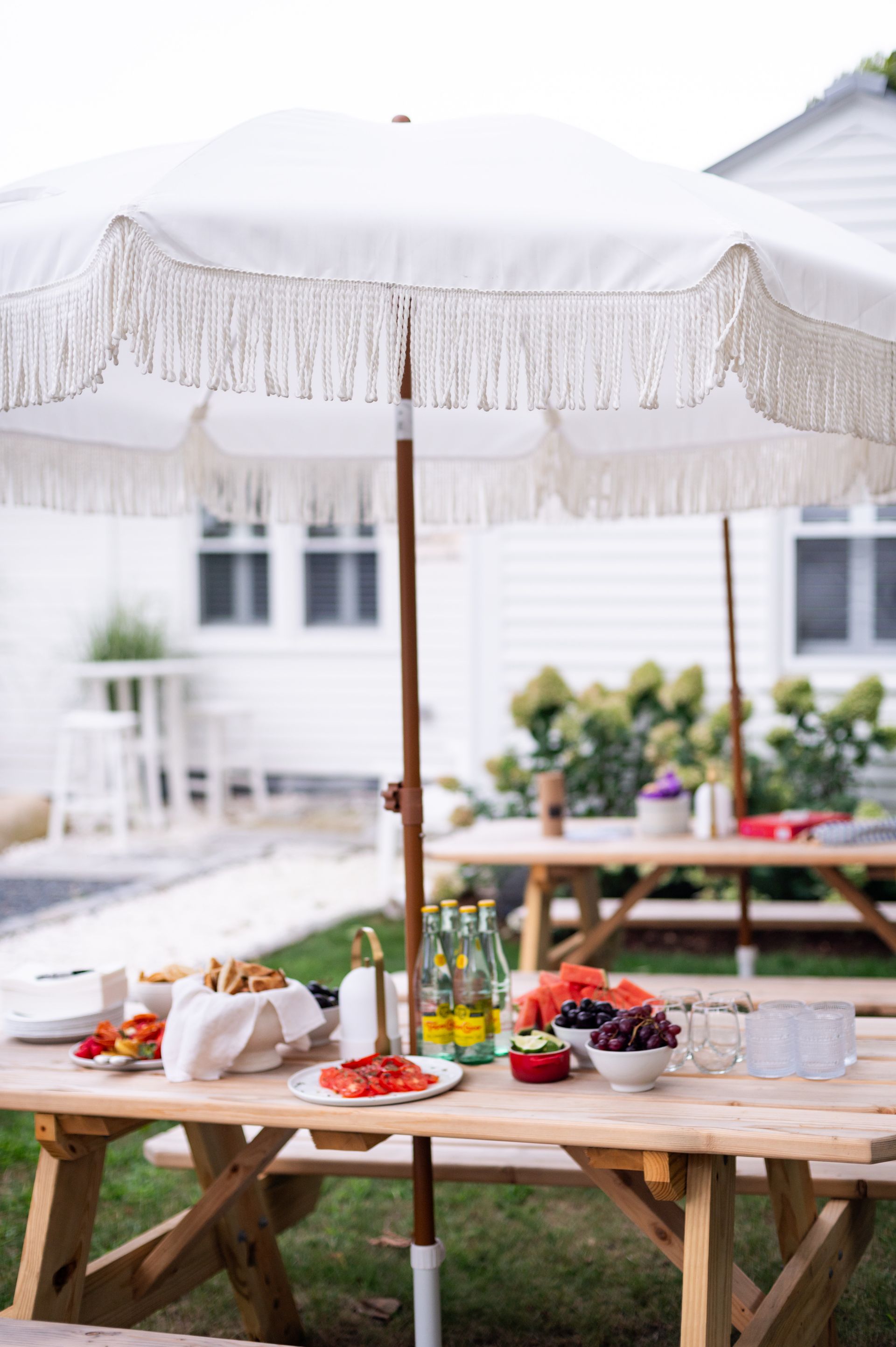 Picnic table set for dining under a white fringed umbrella in a backyard.