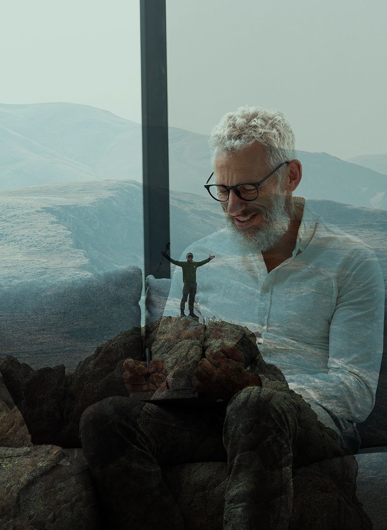 Man with gray hair smiles, holding a small figure on a rock, mountains in the background.
