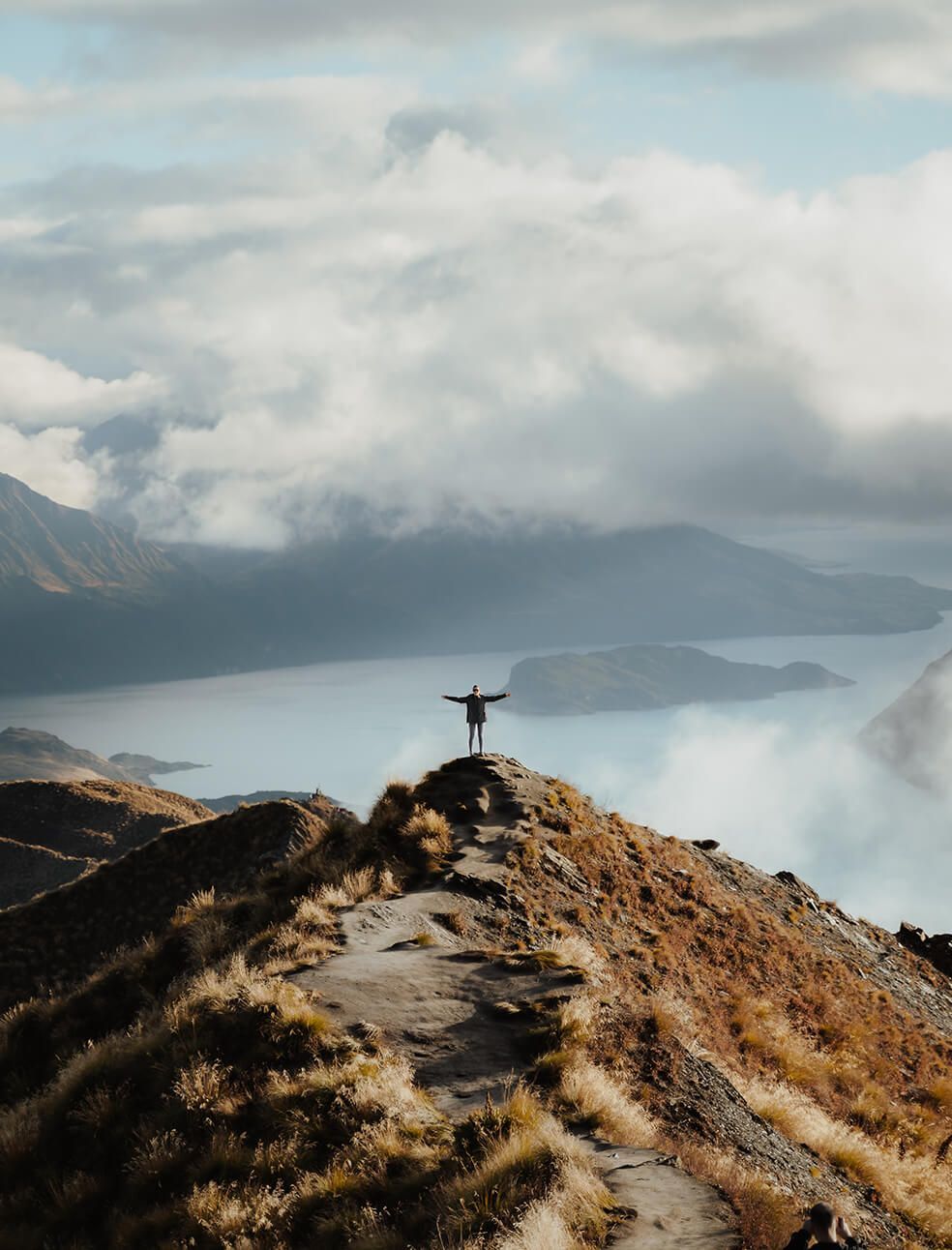 Person with arms raised on a mountain ridge overlooking a lake, cloudy sky.