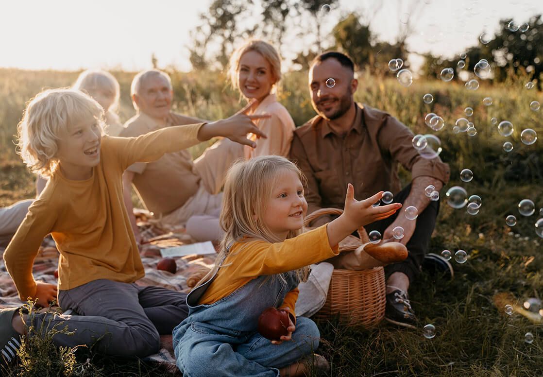 Family picnic outdoors. Two children reaching for bubbles, smiling. Adults seated, smiling, in a field at sunset.