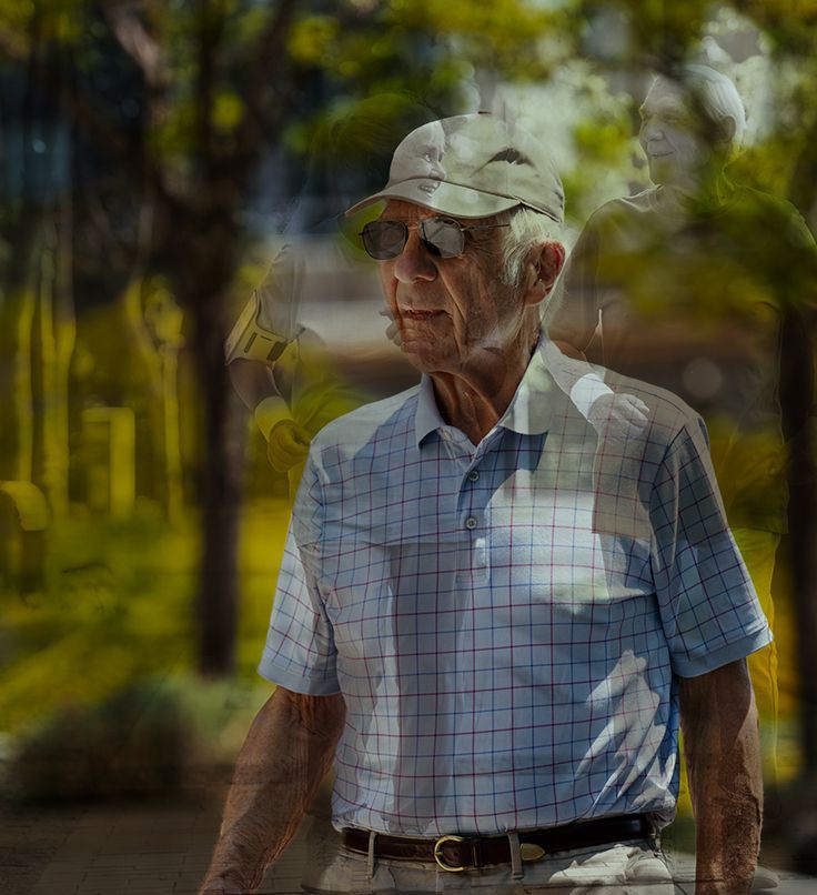Older man in a blue shirt and cap walks outside, wearing sunglasses, with blurred figures and greenery in the background.