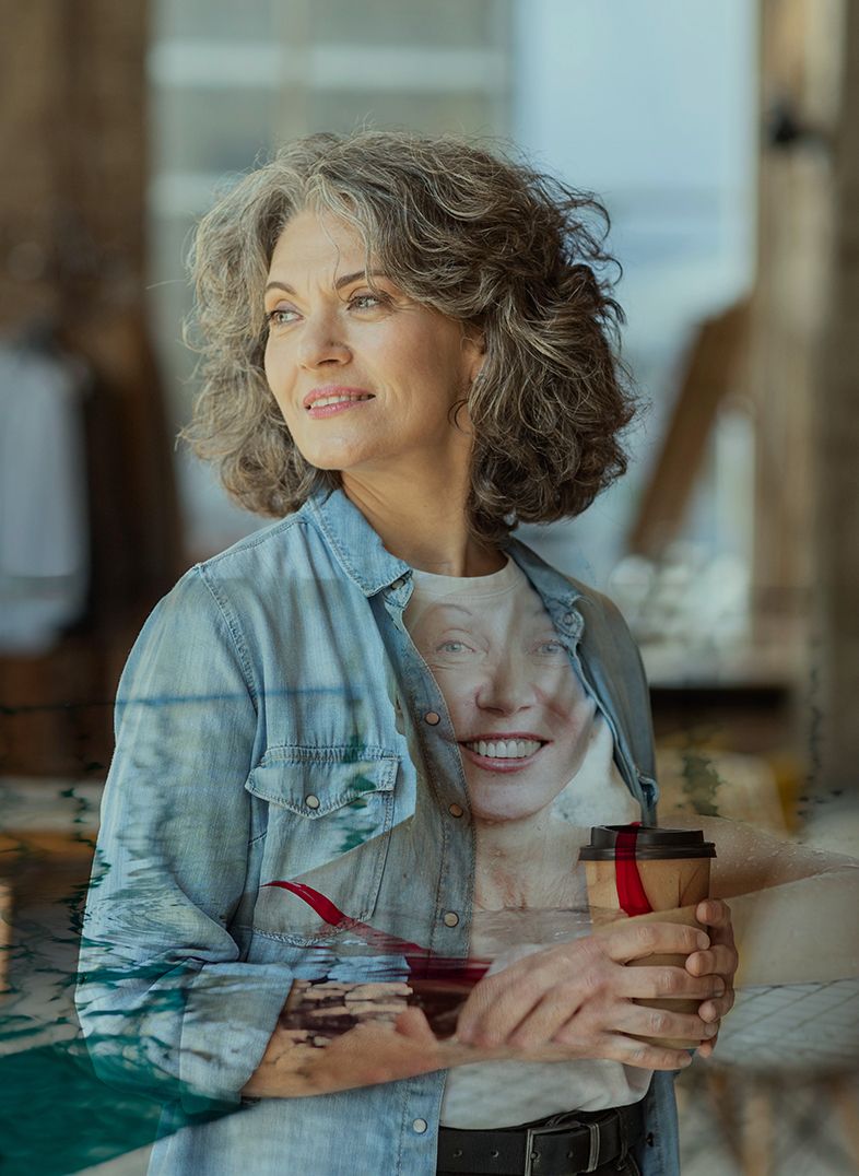 Woman with gray hair, holding a coffee cup, smiles near a window.