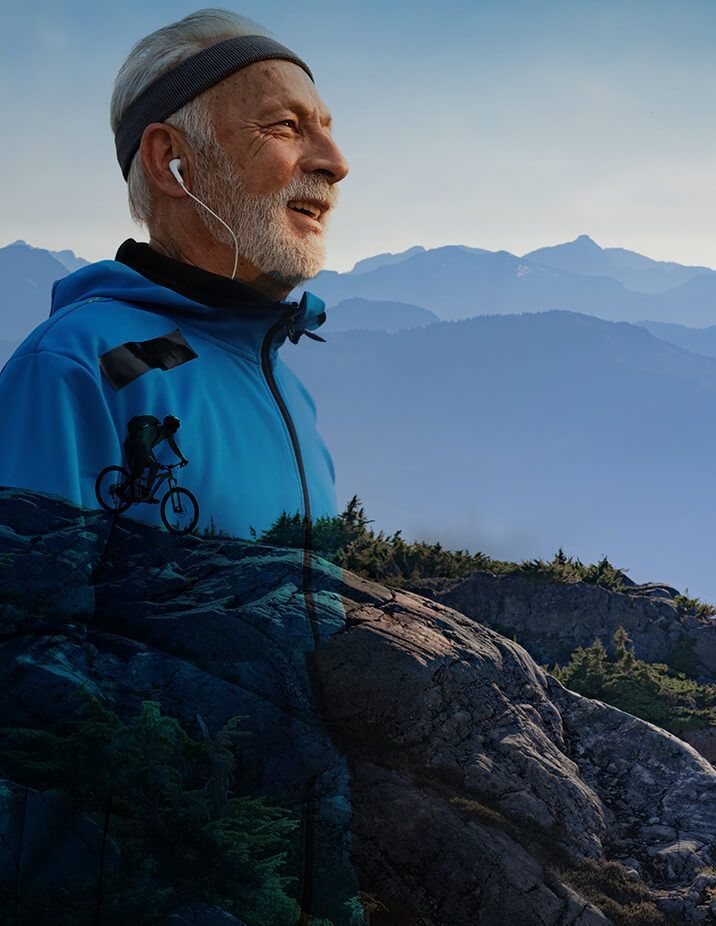 Older man smiling, wearing headphones and blue jacket, in mountains, partially overlaid with a mountain biker on a trail.