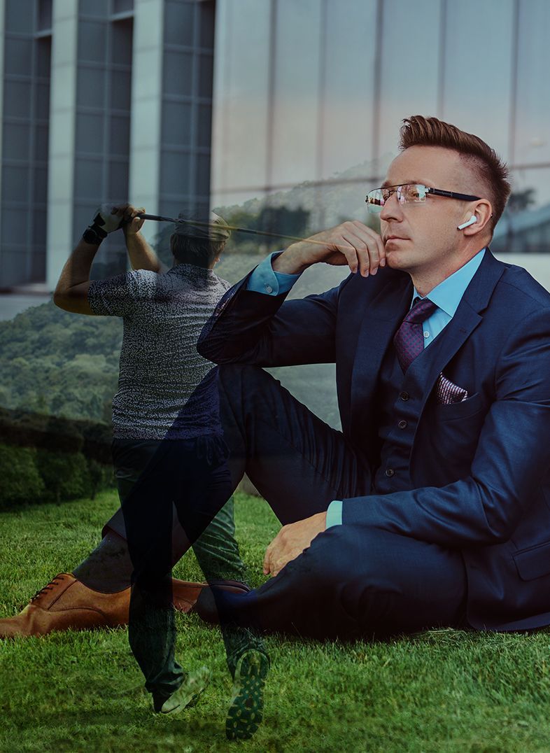 Man in suit sitting on grass with a blurred figure playing golf.