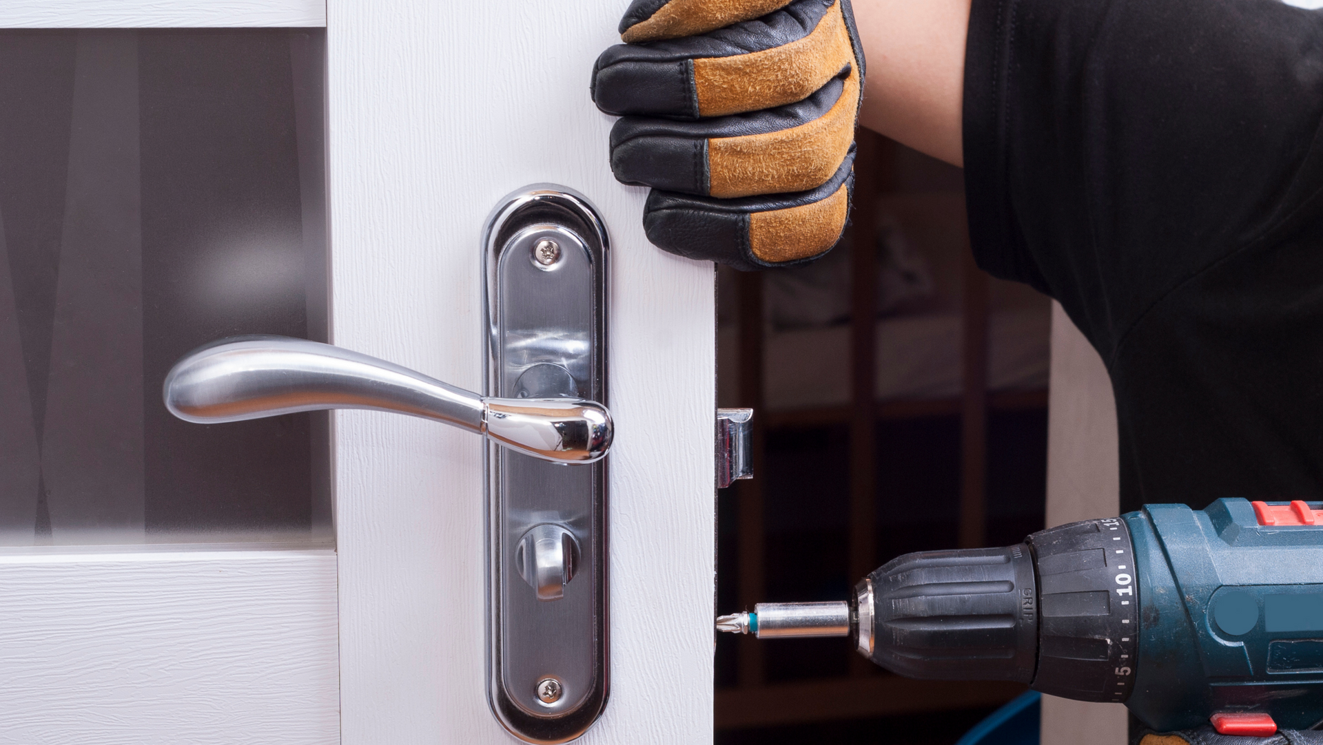 Person with gloved hand using a power drill to install a silver door handle on a white door.