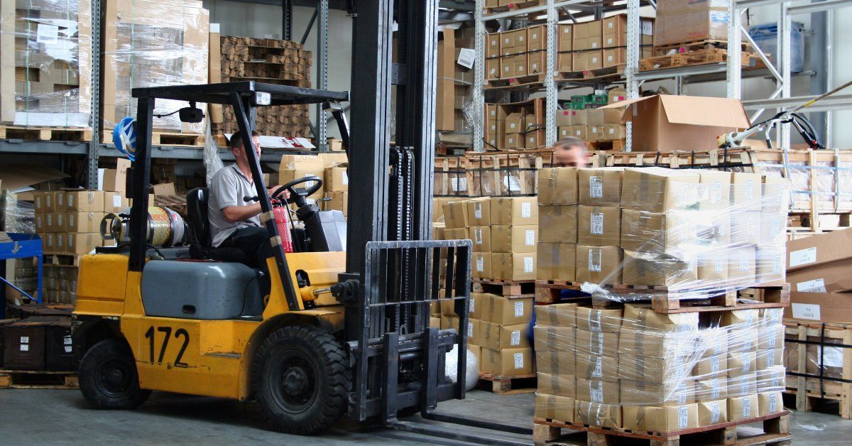 A man wearing a grey shirt is operating a large piece of machinery inside a warehouse filled with boxes.