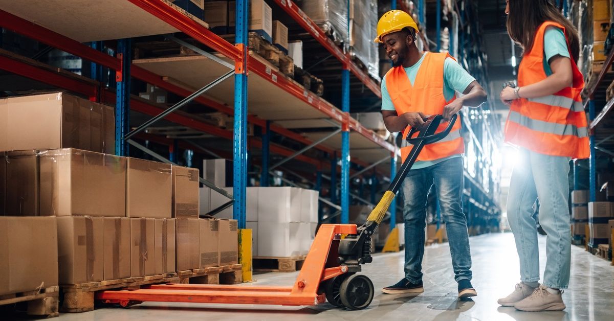 Two workers wearing orange high-visibility vests and yellow hard hats use a pallet jack.