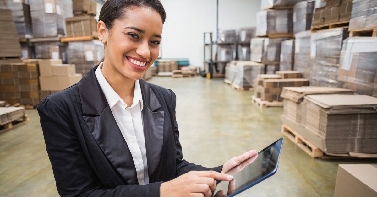 A woman wearing a black blazer smiles as she holds a tablet in a warehouse space filled with flattened cardboard boxes.
