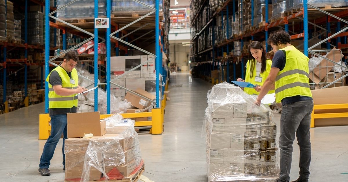 Three people wearing yellow high-visibility vests examine boxes stacked on pallets inside a warehouse.
