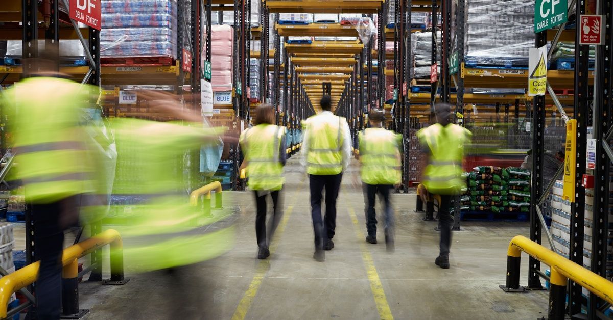 A group of several people wearing high-visibility vests walks down the aisle of a warehouse with several shelves.