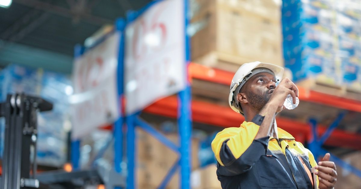 A close-up showing a man wearing a hard hat and jacket drinking out of a water bottle in what appears to be a warehouse.