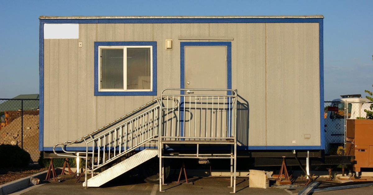 A gray modular room with blue trim. It is outside, the sky above is blue with no cluds. There is a fence behind the room.