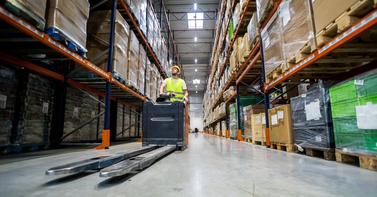 A person wearing a yellow hard hat and hi-vis vest drives a forklift through the wide aisle of a warehouse.