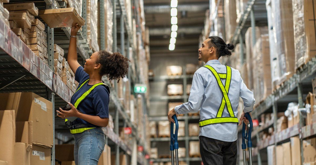 A young woman in a hi-vis vest is picking a cardboard box off a tall shelf.