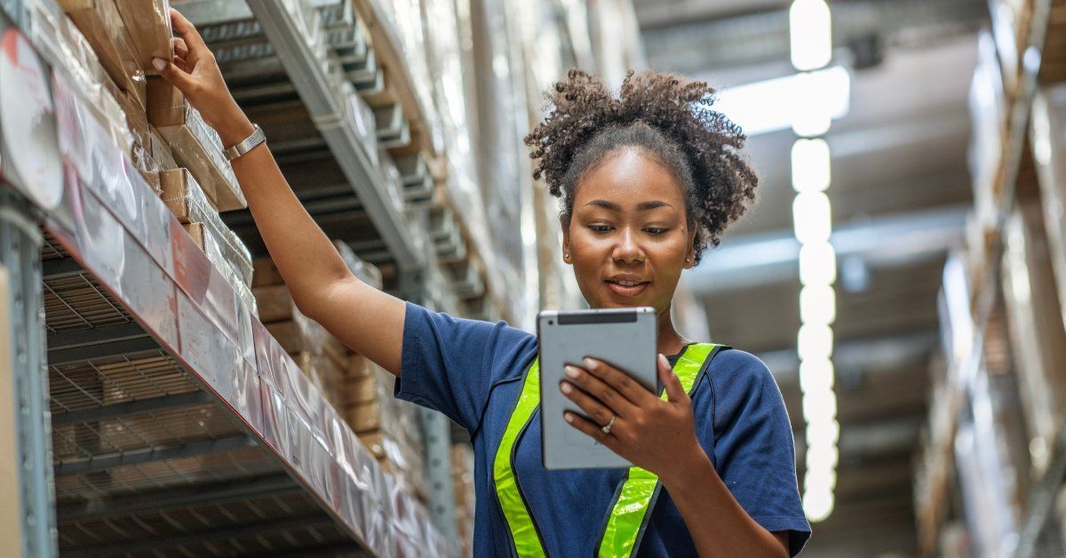 A close-up of a young woman looking closely at a silver tablet as she grabs a cardboard box from a large shelf.