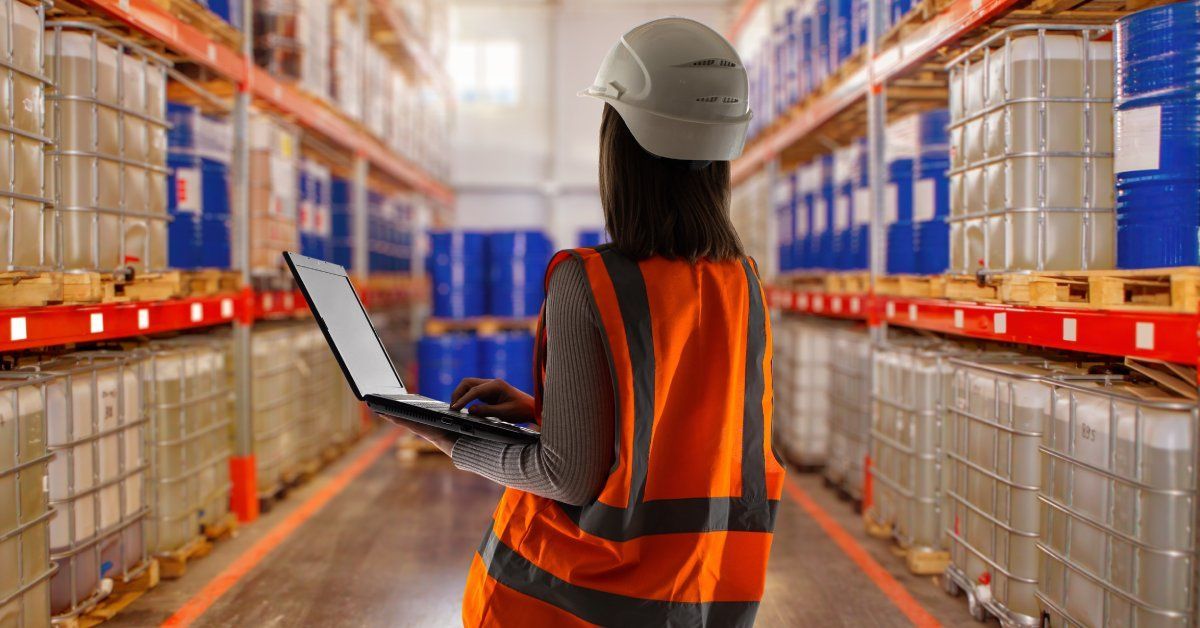 A woman wearing an orange safety vest looks across a large warehouse full of products. She holds a laptop in her hands.