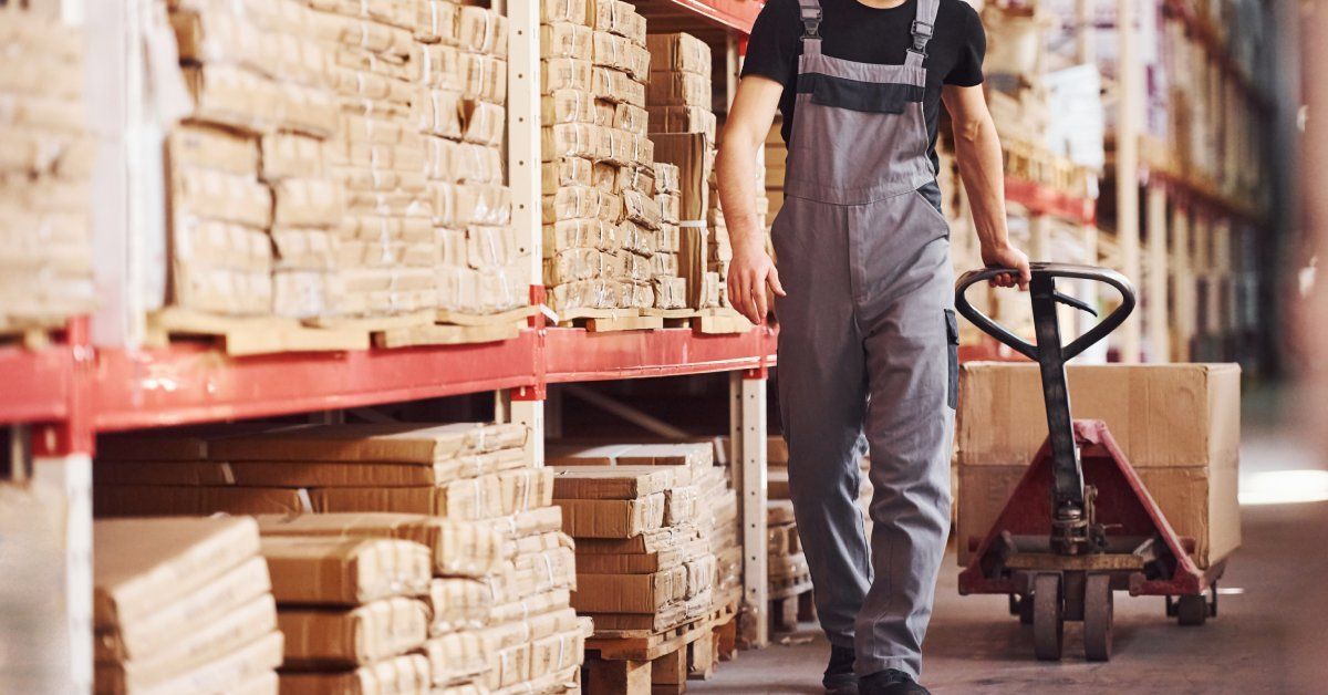 A warehouse worker wearing a hardhat pulls a dolly with a box on it next to large shelving units with other boxes.