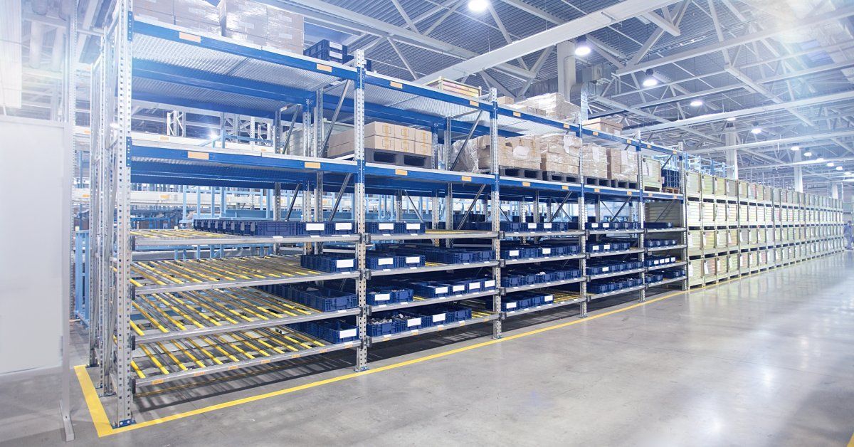 The interior of a brightly lit warehouse. There is a long row of blue shelving units without anything on them.