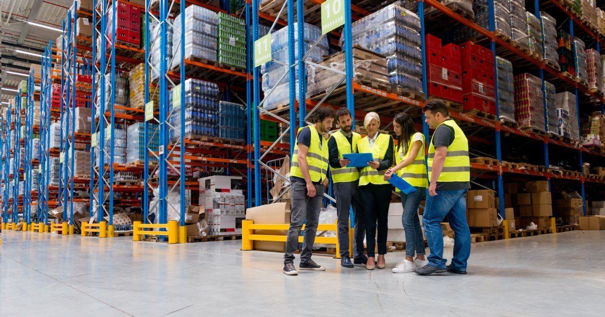 Five people wearing yellow high-visibility vests stand in a warehouse with large shelves while looking at a blue clipboard.