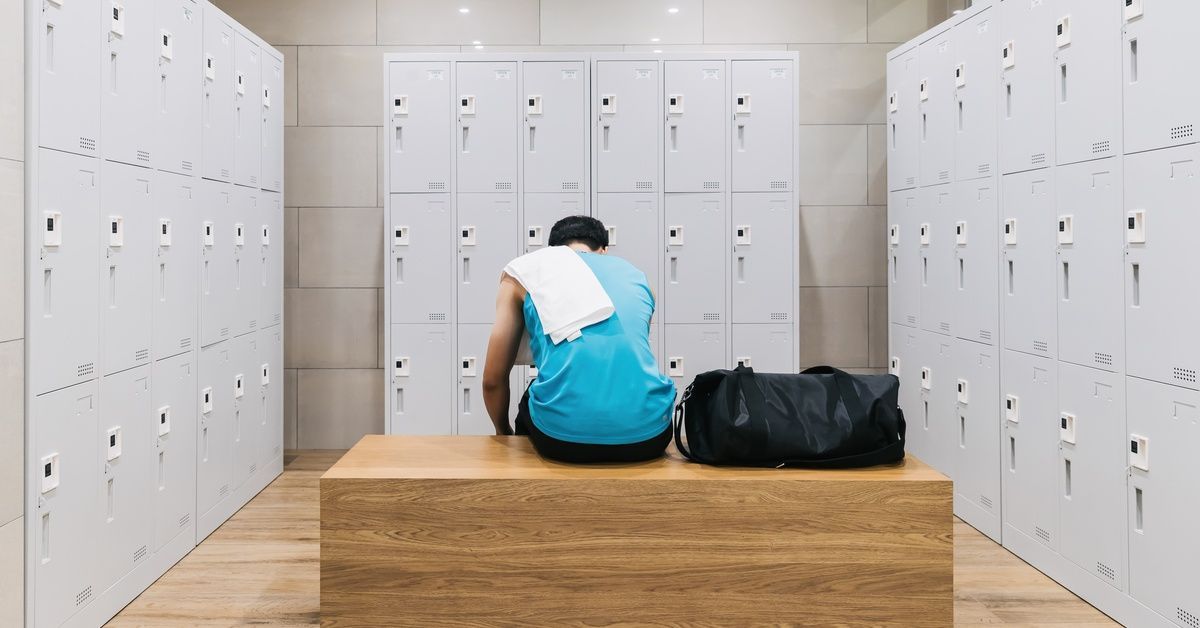 A person with a white towel on their shoulder is sitting on a wooden bench in a room with several gray lockers.