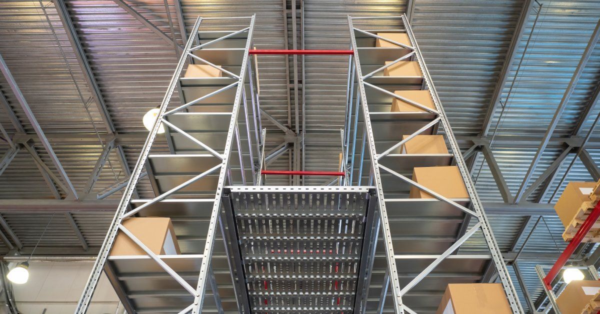 An extreme close-up of a multilevel, metal storage rack that's inside a large warehouse. Cardboard boxes are on the shelves.