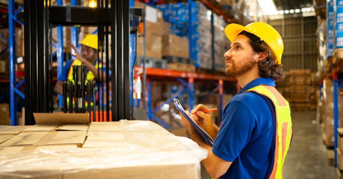 A young man is wearing a high-visibility vest and a hard hat. He's holding a clipboard and inspecting product inventory.