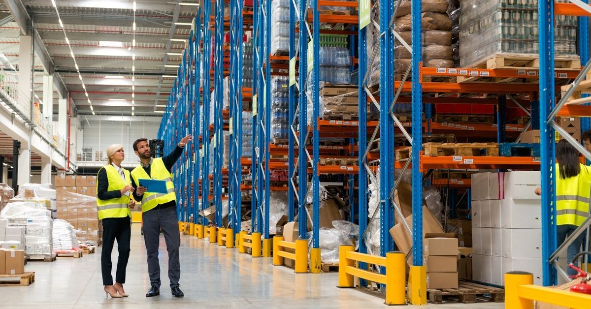 Two managers standing in front of multiple rows of pallet rack shelving. One of them is pointing at something.